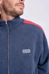 Campera deportiva con cierre frontal, cuello alto y puños y cintura acanalados. Color azul marino con efecto desgastado y dos franjas rojas a lo largo de las mangas. Presenta un pequeño logo bordado en el pecho.