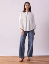 Blusa blanca de gasa con cuello a la base, escote en V y abertura central con botones. Tiene mangas 3/4 amplias con apliques de crochet y detalles plisados en la parte delantera.