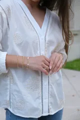 Blusa blanca de manga larga con elástico en puños, escote en pico y cierre frontal con botones. Detalles de bordados con hilado en tonalidad camel y acabado con entredoses a tono.