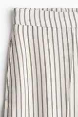 Pantalón ancho de tiro alto con elástico en la parte posterior de la cintura y cierre oculto con corchete en un lado. Bolsillos laterales discretos y piernas amplias con pliegues adelante y atrás. Diseño a rayas verticales grises y blancas.