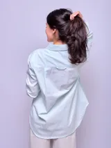 Camisa blanca de algodón, corte holgado, con cuello clásico y botones frontales. Las mangas son largas y se ven remangadas.