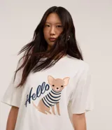 Remera blanca de algodón BCI con cuello redondo y mangas cortas. Presenta un bordado de un chihuahua con un suéter a rayas y la palabra "Hello".