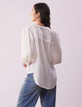 Blusa color beige de gasa con cuello a la base, escote en V y abertura central con botones. Presenta detalle de crochet en hombros y espalda. Manga 3/4 amplia con puño y botón.