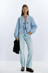 Camisa celeste con rayas verticales marrones y blancas, cuello redondo con escote en pico y mangas largas abullonadas. Cierre frontal con lazos con pespuntes marcados.