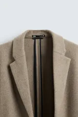 Abrigo straight fit color beige confeccionado en tejido con lana. Cuello con solapas de muesca, manga larga acabada en puño con botones, bolsillos de vivo en cadera, detalle de bolsillo interior y bajo con abertura en centro espalda. Cierre frontal de botonadura.
