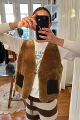 Chaleco marrón de cuero con interior de corderito color crema, cierre con gancho metálico y bolsillos frontales de corderito.