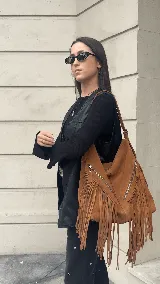 Bolso tipo hobo color tostado, hecho de cuero con flecos largos en el frente y dos bolsillos con cierre. Tiene una manija regulable con hebillas metálicas.