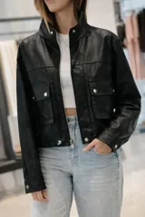 Campera corta de símil cuero negra, con cuello alto abotonado, cierre frontal metálico y dos bolsillos frontales con tapa y broche. Presenta un calce relajado y puños con broche.