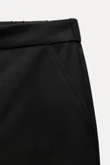 Pantalón tobillero negro de tiro medio con cintura elástica en la espalda, bolsillos delanteros y bolsillos de parche con solapa en la parte trasera. Bajo acabado con pinzas. Cierre frontal con cremallera y botón.