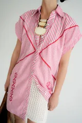 Vestido camisero de algodón a rayas verticales rojas y blancas, con bordados florales rojos en el frente y en la falda. Presenta cuello camisero, cierre frontal con botones y mangas cortas.