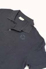 Polo de manga corta color azul oscuro, con cuello y tapeta de botones negros. Presenta un pequeño bordado circular en tono azul claro en el pecho.