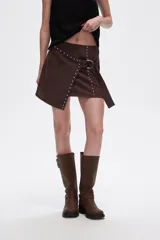 Minifalda marrón de gamuza sintética con corte irregular, detalle de tachas metálicas y cinturón con hebilla redonda.
