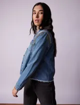 Campera de jean celeste de la marca Baccini, con cierre central, bolsillos delanteros, apliques colgantes de tachas en los hombros y terminación desflecada en el ruedo.