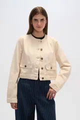Chaqueta corta color crema de algodón, con cuello redondo, cierre frontal con botones dorados, mangas largas y dos bolsillos de parche en el pecho.