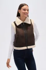 Chaleco sin mangas de eco-cuero marrón con forro y cuello de piel sintética color crema. Presenta cierre frontal y detalles de hebillas ajustables en los laterales.