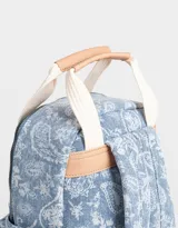 Mochila de denim azul con estampado paisley blanco, bolsillo frontal con cierre, correas regulables y manijas superiores de algodón con broche de cuerina. Acceso mediante cierre superior y puller de cuerina. Incluye tag de cuerina.