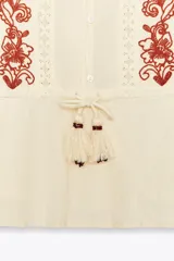 Blusa color crema de cuello redondo y manga larga con elástico en los puños. Presenta bordados florales en el frente y mangas, puntilla a tono y cintura ajustable con cordón y borlas. Cierre frontal con botones.