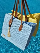 Bolso de playa tejido con trama abierta, color celeste, con forro interior y asas de cuero marrón. Incluye un dije con cuentas de madera y borla.