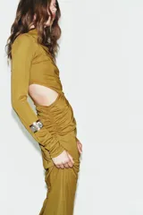 Vestido largo color mostaza con cuello alto, manga larga drapeada y detalles cut out laterales. Presenta frunces en la cintura y la espalda, y bajo recto.