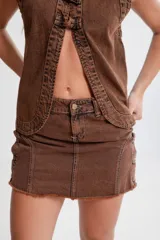 Minifalda marrón de denim con efecto desgastado, corte ajustado, cintura baja, cierre frontal con botón metálico y ruedo desflecado. Presenta botones metálicos decorativos en los laterales.