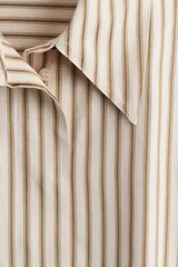 Camisa oversized de sarga color beige con rayas verticales marrones, cuello clásico, cierre frontal con botones ocultos, mangas largas con puños abotonados y bajo recto.