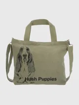Bolso tipo tote de lona color verde oliva con doble asa de mano y correa bandolera ajustable y extraíble. Estampado frontal con un perro basset hound y el logo de la marca Hush Puppies.