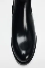Botín Chelsea de cuero negro con elásticos laterales, puntera redondeada y detalle de placa metálica dorada en el tacón.