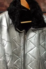 Chaqueta de cuero plateada con acabado metalizado y diseño acolchado. Presenta cuello y puños de piel en color negro, cierre frontal metálico y bolsillos laterales con cremallera.
