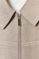 Chaqueta corta color beige con cuello solapa, manga larga y cierre frontal con cremallera.