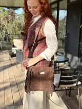 Cartera bandolera de cuero color chocolate, con forro interno de tela y flecos en la parte inferior. Tiene asa larga regulable y cierre superior con botón. Fabricada en Uruguay.