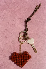 Llavero con cordón marrón y blanco, llave dorada y dije colgante de corazón rojo hecho con cuentas. Incluye aros metálicos dorados.