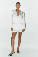 Blazer oversize de satén color blanco, con cuello solapa, escote pico cruzado, manga larga con hombreras, bolsillos de doble vivo y abertura en la espalda. Cierre frontal cruzado con botones forrados.