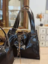Bolso tipo tote de cuero negro brillante con doble asa de hombro y cierre con cordones.
