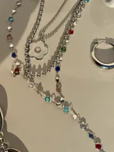 Set de tres collares plateados de diferentes largos. El más corto es tipo choker, el segundo tiene un dije de flor y el más largo es tipo corbatín con piedras de colores.
