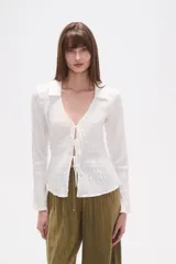 Blusa blanca de algodón con textura fruncida, manga larga y escote en V con lazo ajustable en el centro del busto.