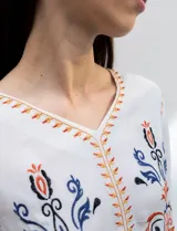 Blusa blanca de rayón con escote en V y mangas largas. Presenta bordados florales en tonos naranja, azul y negro en la parte delantera y en las mangas.