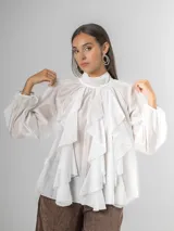 Camisa blanca de algodón italiano con cuello alto ajustable mediante lazo trasero, mangas abullonadas y un gran shabot frontal con volados en cascada.