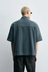 Camisa de denim de algodón, color beige claro, con corte relajado, cuello bowling y manga corta. Presenta cierre frontal con botones a presión y un pequeño bordado a tono en el pecho.