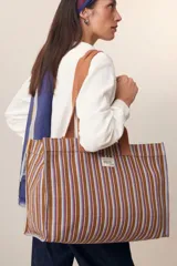 Bolso tote de algodón a rayas verticales marrones, azules y rojas, con asas de mano marrones. Tiene un bolsillo lateral interior grande, un bolsillo para el teléfono y un bolsillo para las llaves.