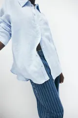 Camisa celeste de lino liviano, con cuello, botones al frente, canesú de doble capa en la espalda, hombros caídos, mangas largas con puños abotonados, bolsillo superior de parche y bajo levemente redondeado.
