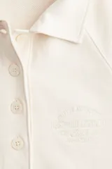 Buzo color crema de algodón con cuello camisero, tapeta de botones y mangas raglán. Tiene un pequeño bordado en el pecho y ribete desflecado en puños y bajo.