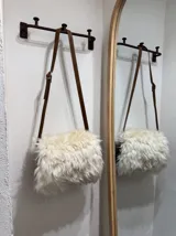 Bandolera de cuero color negro con forro interior y cierre. Presenta un cuerpo cubierto de lana de oveja color blanco y correa larga de cuero marrón con hebilla ajustable.