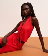 Vestido midi rojo, con textura plisada o arrugada, escote halter en V y detalle de amarras ajustables en el centro del busto que generan una abertura frontal. La falda presenta un fruncido lateral y una abertura alta.