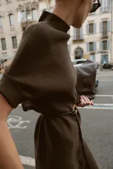 Vestido negro de tejido de punto, corte midi y silueta holgada, con cuello alto acanalado y mangas cortas tipo murciélago. Se ajusta a la cintura con un cinturón fino de efecto piel negro con hebilla dorada en forma de C.