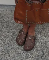 Zuecos marrones de cuero con hebilla metálica y detalles de bordado artesanal, tachas y remaches.