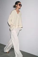 Camisa color crudo de cuello redondo con escote pico y lazada. Manga larga acabada en volantes. Detalle de bordados combinados a contraste y puntilla a tono.