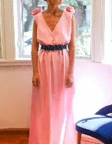 Vestido largo color rosa pálido, con escote en V cruzado, sin mangas y cinturón negro con apliques de flores. Presenta detalle de volados en los hombros y corte evasé.