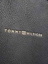 Mochila negra de cuero sintético con textura granulada, cierre de cremallera, asa superior con cinta con el logo de Tommy Hilfiger, correas ajustables, bolsillo interior con cremallera, bolsillo exterior con cremallera y detalles metálicos dorados.