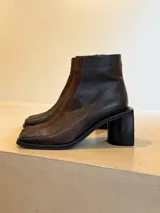 Botinetas de caña media con diseño bicolor en tonos oscuros, combinando paneles de cuero texturizado con detalles en gamuza. Presentan punta cuadrada y taco bloque robusto.