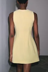 Vestido corto color amarillo pastel, de cuello redondo y sin mangas. Presenta pliegues con volumen en el ruedo y cierre trasero con cremallera oculta.
